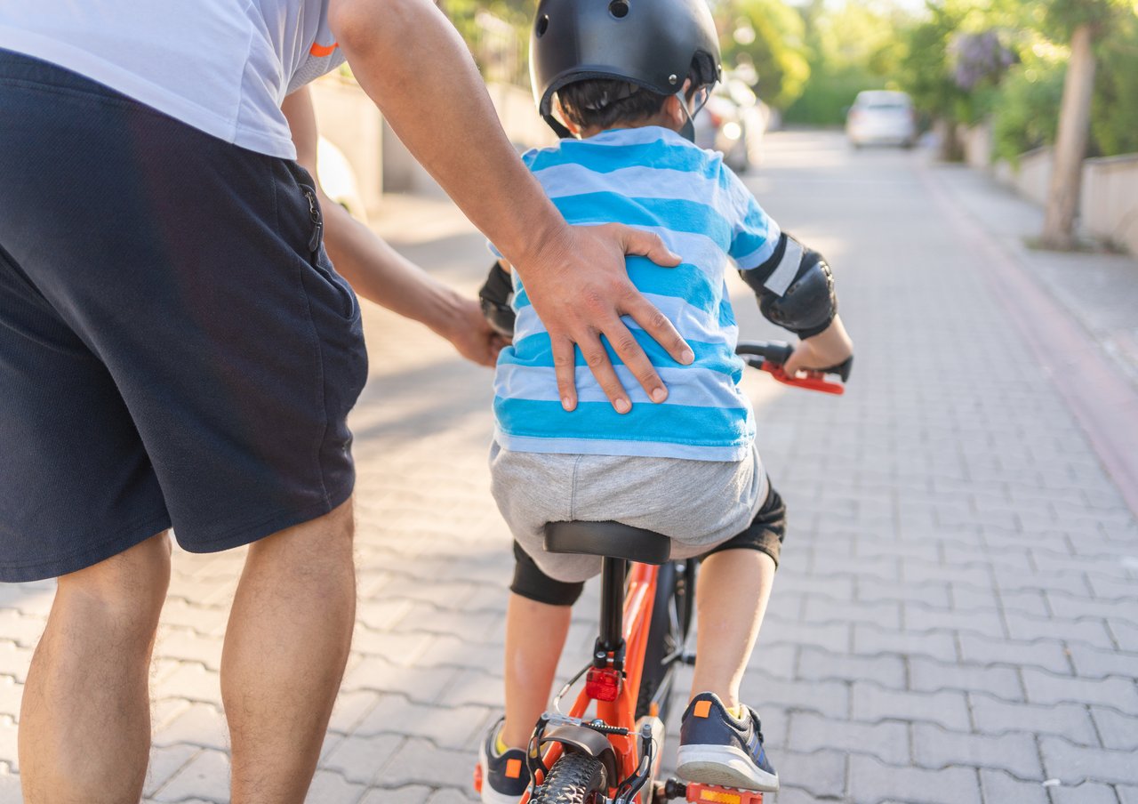 Ein Erwachsener stützt mit der Hand den Rücken eines Jungen mit Helm und Schutzausrüstung, der auf einem orangefarbenen Fahrrad eine gepflasterte Straße entlangfährt.